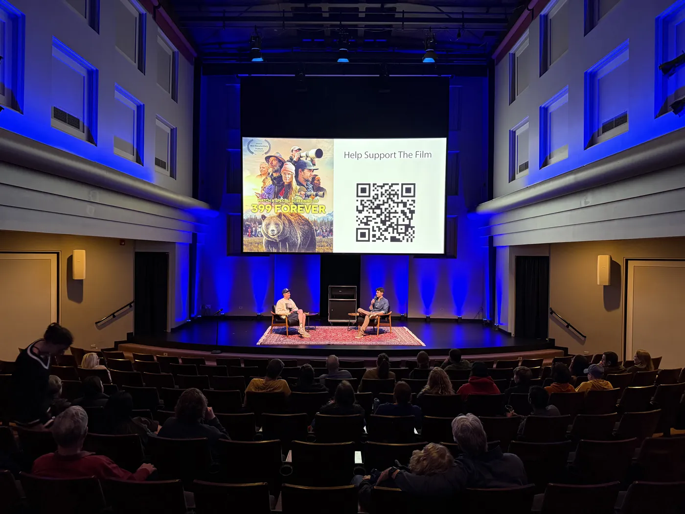 A theater lit in blue with two speakers on stage in front of a 399 Forever slide
